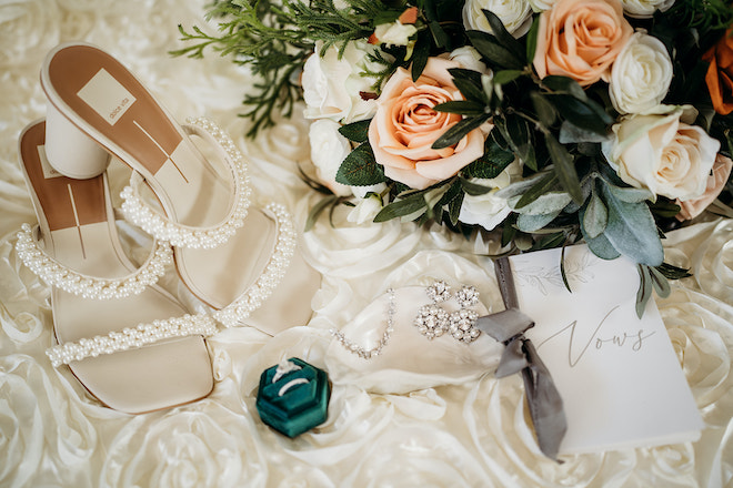The brides shoes, jewelry, bouquet and vows laying in a white rose background.