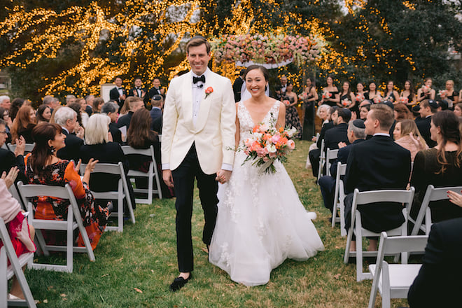 The bride and groom smiling walking down the aisle with the floral-filled chuppah and lit up tree behind them. 