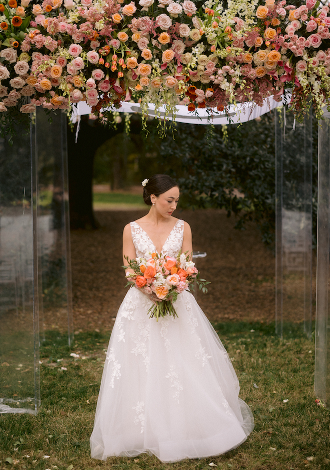 The bride standing under the floral-filled chuppah holding a bouquet of orange and pink florals. 