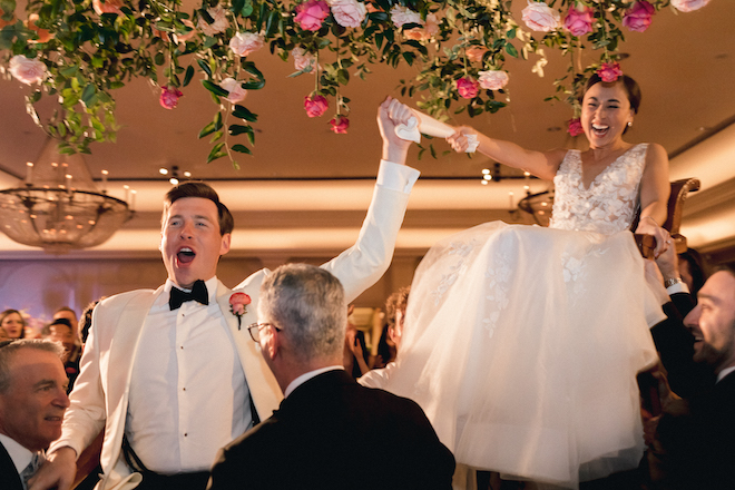 The bride and groom smiling as they are lifted on chairs by their guests.