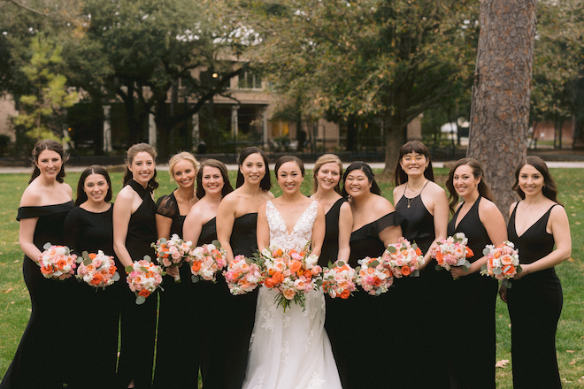 The bride smiling with her bridesmaids wearing black gowns and holding orange, pink and white bouquets. 
