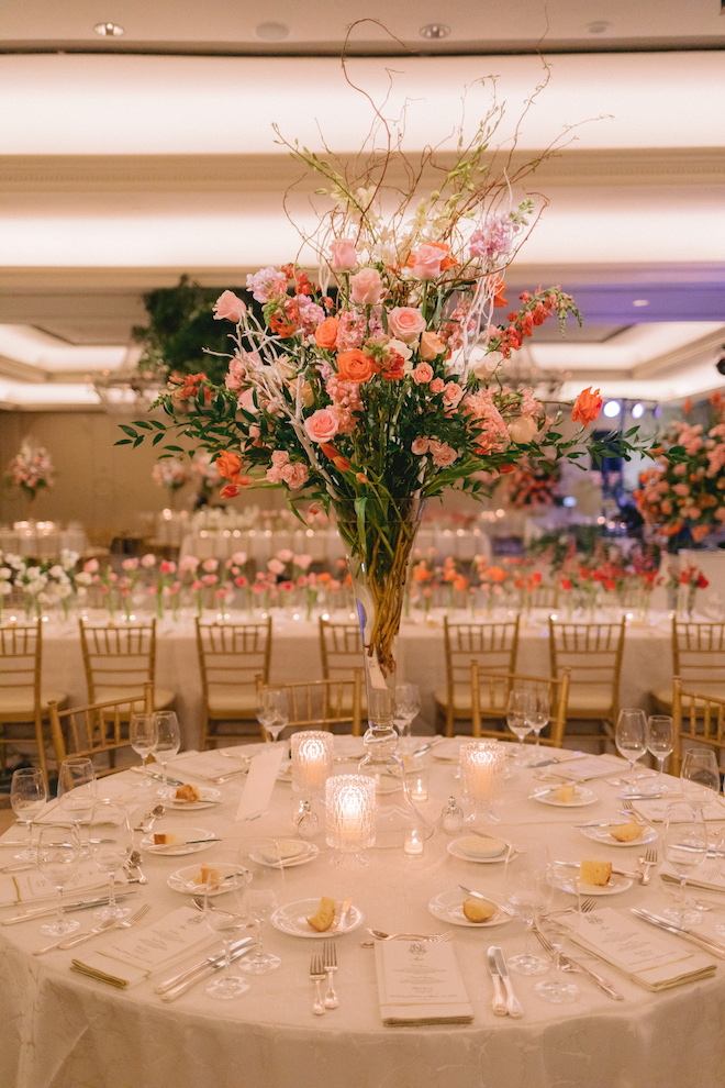 A pink and orange floral centerpiece with greenery and twigs on a white round reception table. 