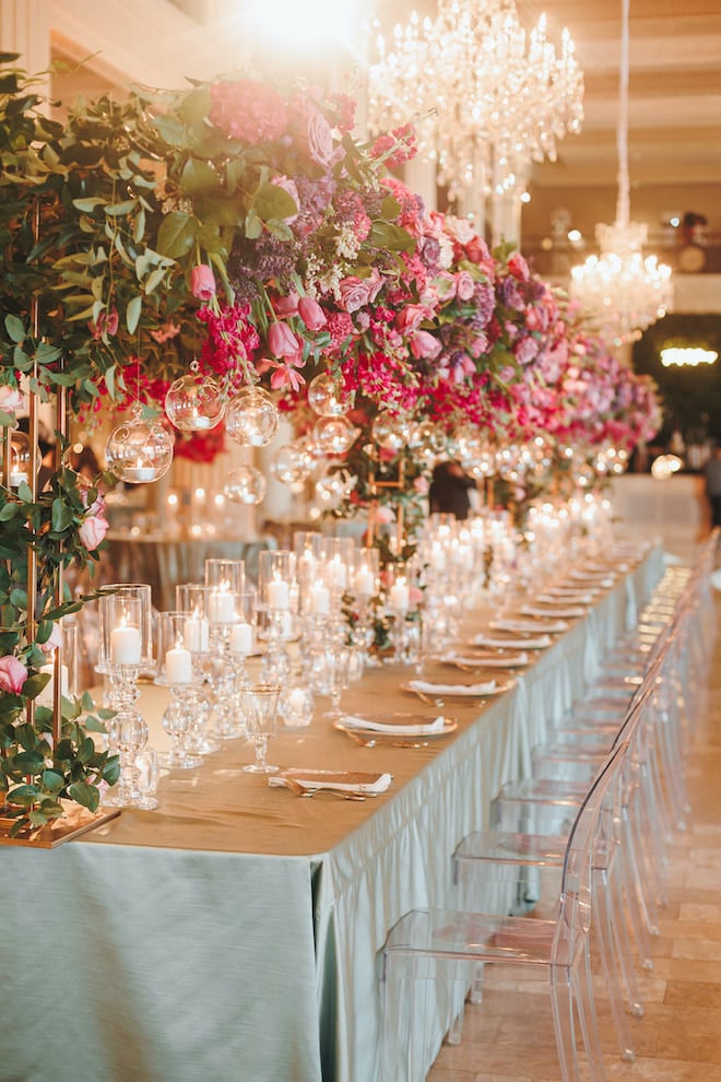 Family style table set with a floral installation of pink and purple flowers, greenery and hanging orbs with lit candles at a wedding reception. 