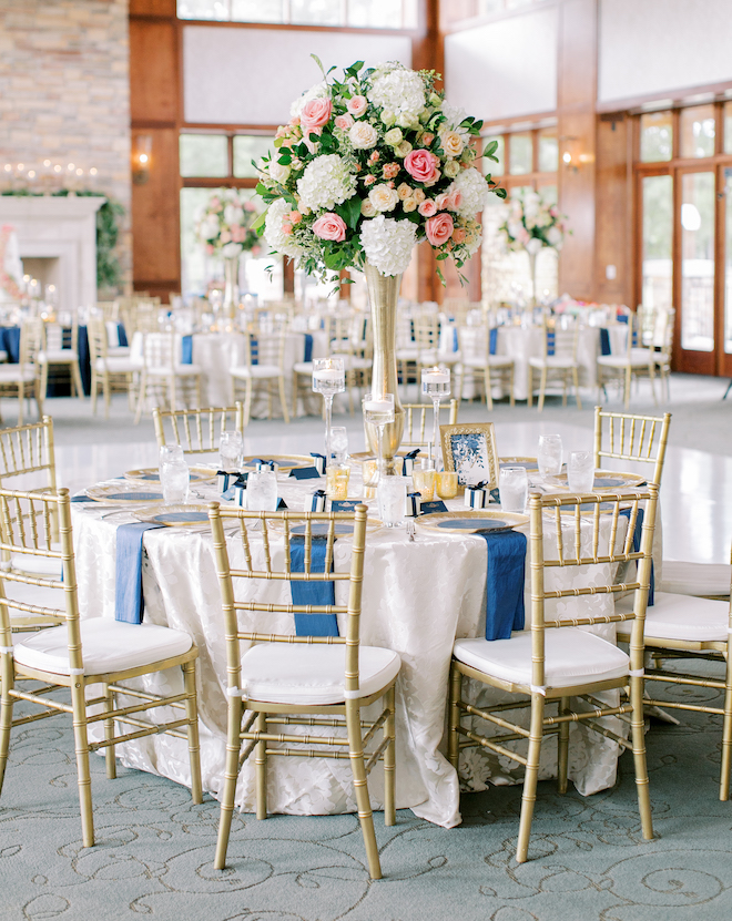 A round table at the reception with gold chairs, blue napkins and pink and white floral centerpieces.