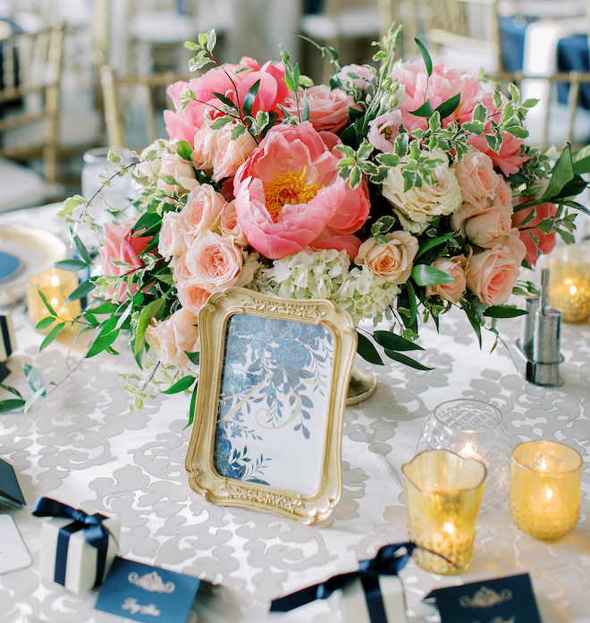 A pink, blush and white floral arrangement on a reception table with a framed blue and gold print with the number "13". Candles and white and blue boxes are at each seat. 