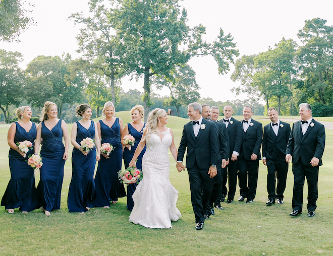 The bride and groom holding hands and walking with the bridesmaids in navy dresses and the groomsmen in black suits walking behind them.