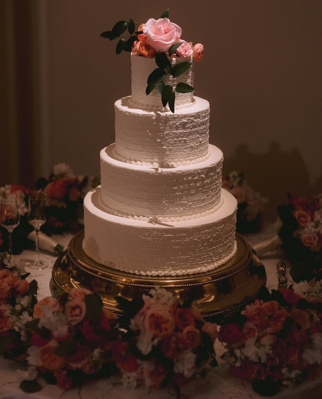A four-tier white wedding cake with pink and orange flowers as the cake topper. 