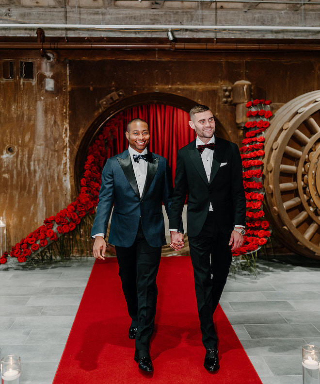The two grooms walk down the red carpet aisle holding hands at The Vault at Corinthian Houston.