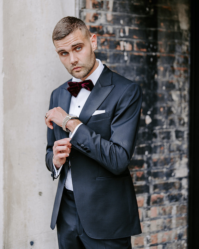 The groom stands outside in his tuxedo with a maroon bowtie outside their wedding venue, The Vault Corinthian Houston.