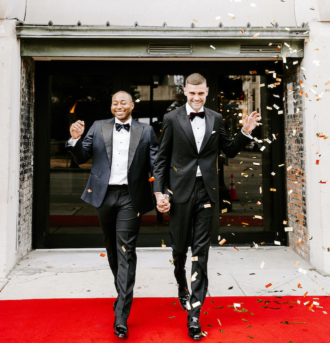 The two grooms exit the wedding venue holding hands with a confetti celebration. 