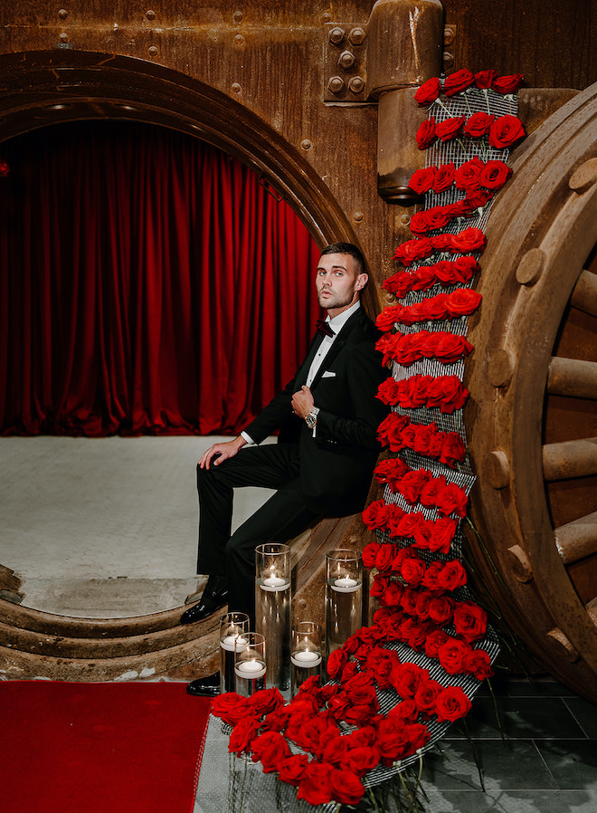 The groom poses up against the vault with red roses and glass pillar candles. 