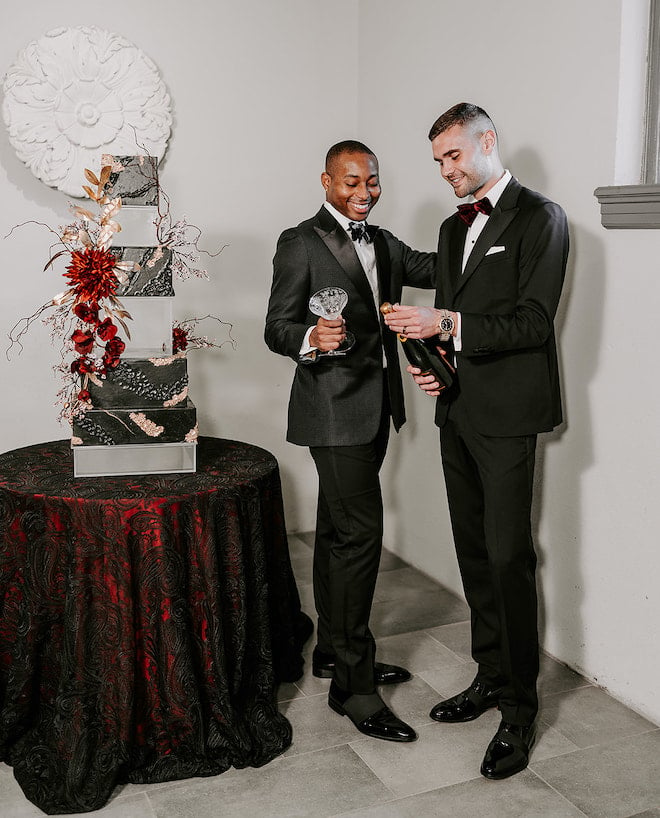 The two grooms open a bottle of champagne next to their wedding cake designed by Susie's Cakes.
