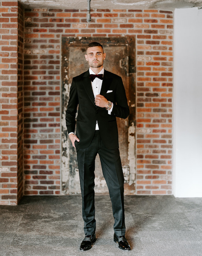 The groom poses in a tuxedo in front of a brick wall at The Vault at Corinthian Houston.