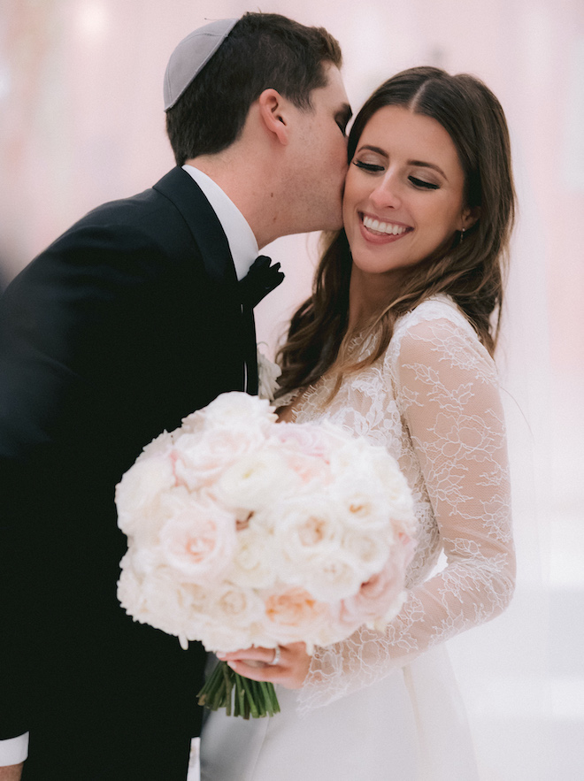 The groom kissing the bride's cheek as she hold her bouquet of roses.
