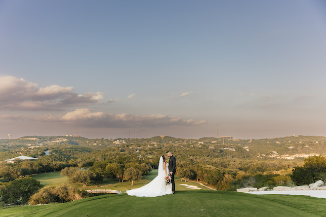 A bride and groom gazing at each other on a lawn with a hill country view.