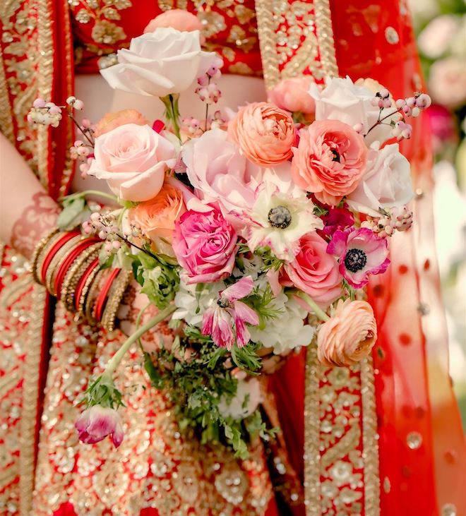A bride wearing a red and gold lehenga holding a bouquet of summer-colored florals in corals, pinks and white. 
