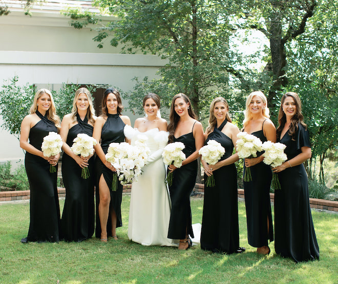 The bride smiling with her bridesmaids in black gowns holding white bouqets.