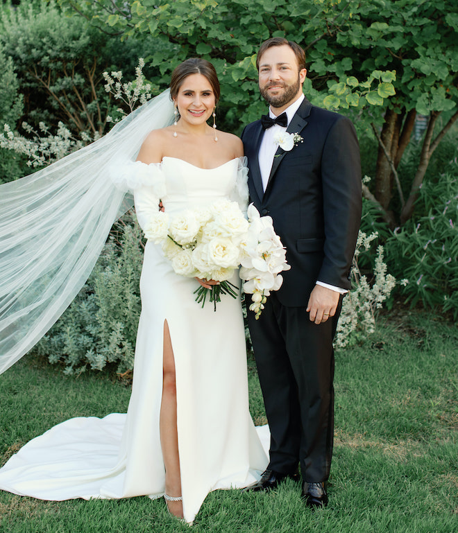 The bride and groom smiling as the bride holds a bouquet of white roses and orchids at their wedding in the San Antonio hill country.