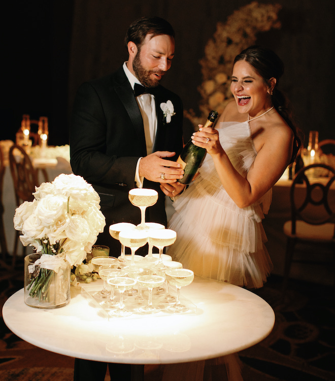 The bride and groom laughing as they pour champagne on a tower of glasses.