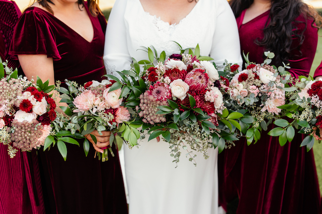 A bride and bridesmaids holding bouquets with greenery, white and maroon florals. 