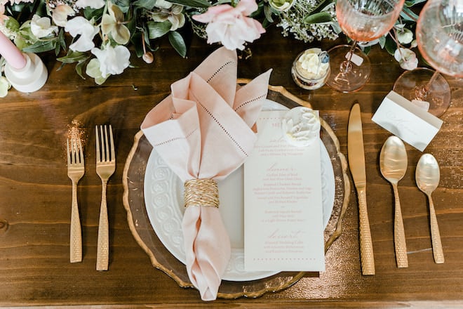 Gold flatware and pink linens with elegant stationery, making for a light and bright tablescape.