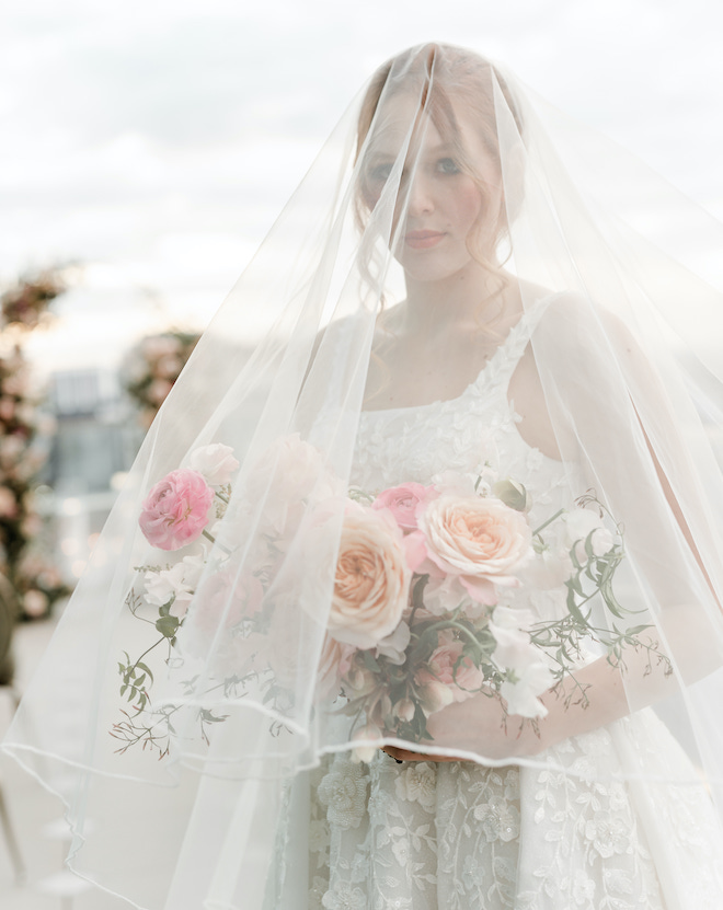 The bride holding a bouquet of light and bright florals with her veil over her head.