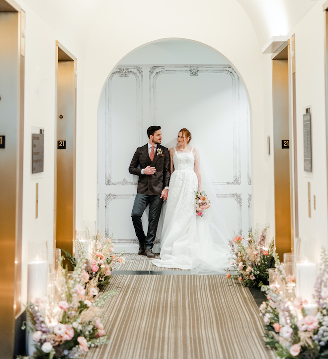 The bride and groom looking at each other at the end of a hallway of elevators.