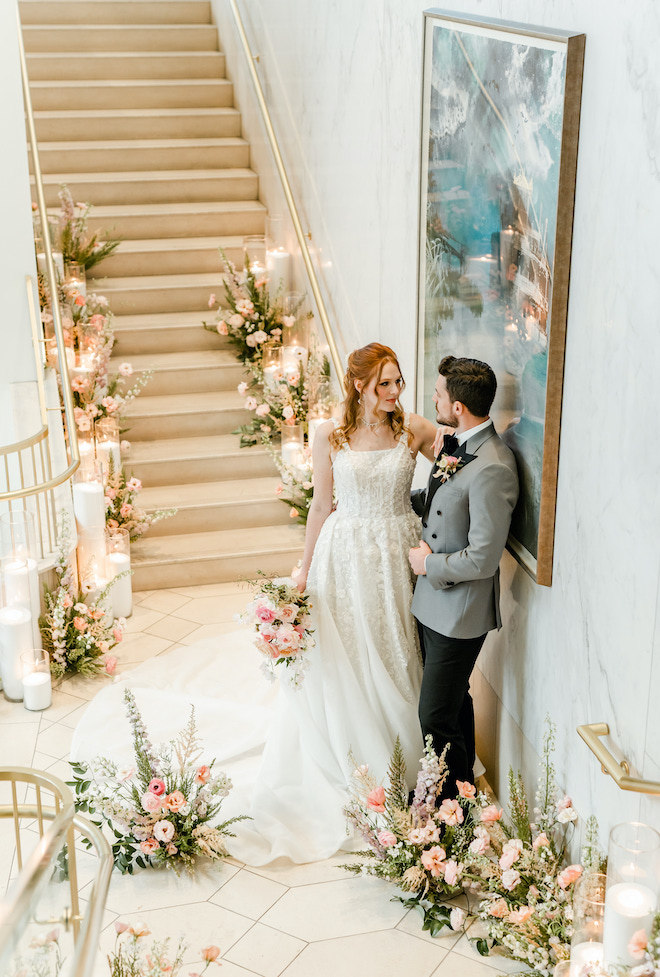 The bride resting her hand on the groom's shoulder on the grand staircase.