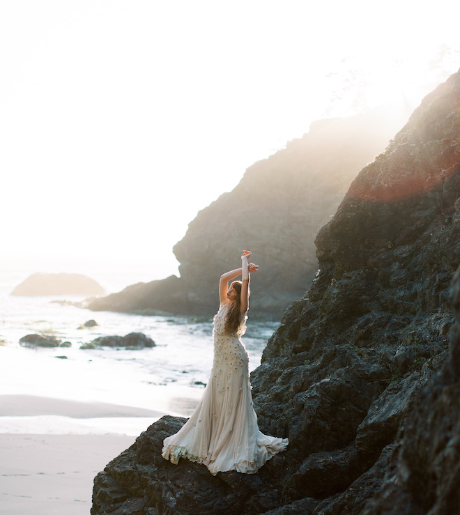 The bride holding her hands above her head while standing on a cliff off of the ocean.