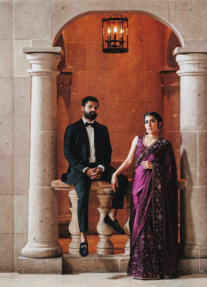 The groom sitting on a ledge while the bride stands with her arm on his knee.