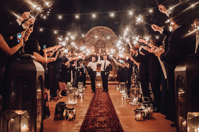 Two grooms walking down the aisle as their guests wave sparklers at Madera Estates.