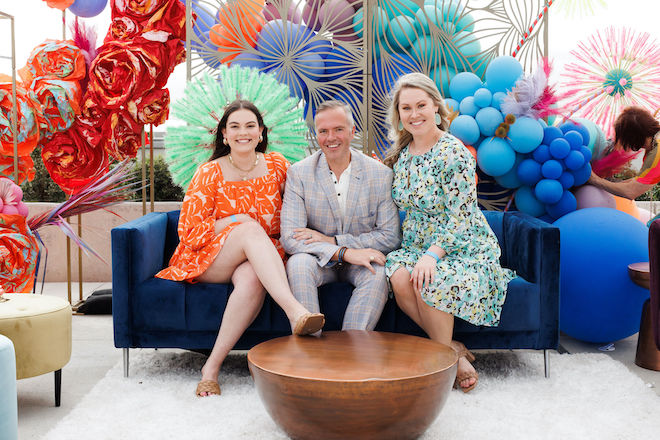 Three wedding professionals sitting on a blue couch with a colorful balloon backdrop.