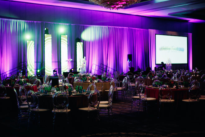 An indoor event space decorated with round tables and ghost chairs, facing a stage with purple lighting.