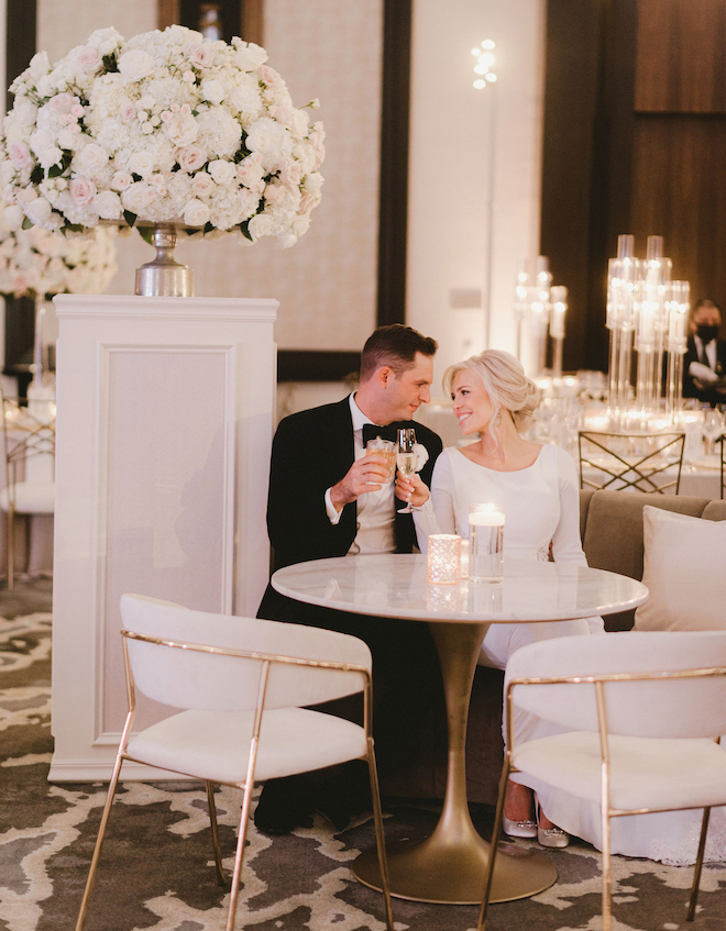 A bride and groom smiling at each other and clinking glasses as they sit in the ballroom of the Post Oak Hotel at Uptown Houston.