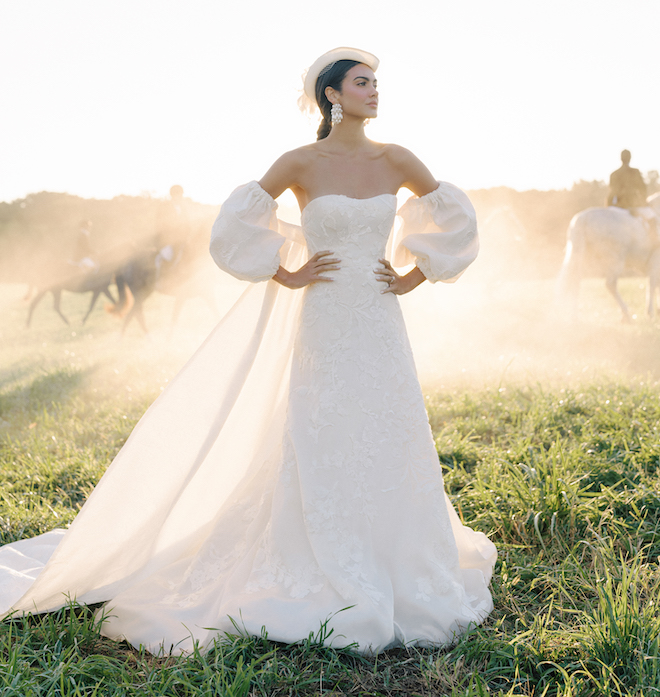 A woman wearing an Anne Barge wedding gown with puffy sleeves a veil and a hat standing in a field with horses behind her. 