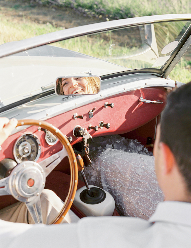 The groom driving the car as the bride smiles through the rear view mirror.