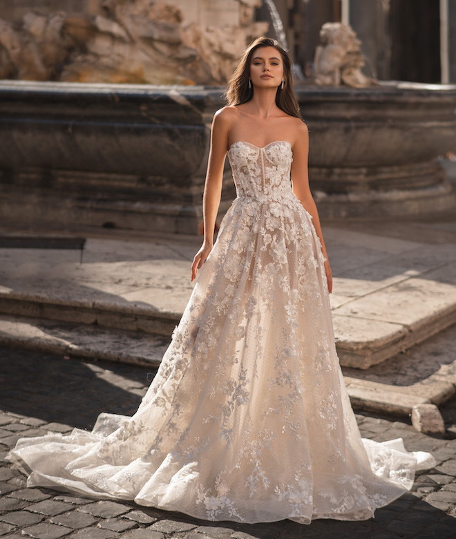 A woman wearing a wedding gown with a corset style top and floral embellishments standing in front of a fountain. 