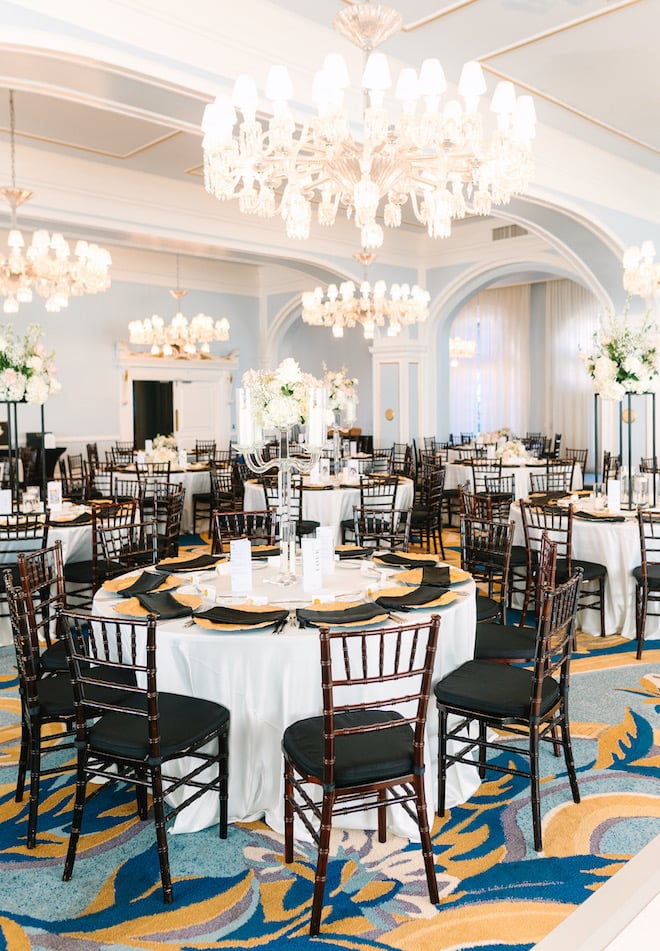 A blue ballroom known as The Music Hall decorated with chandeliers and round tables with white tablecloths and black and gold table settings.