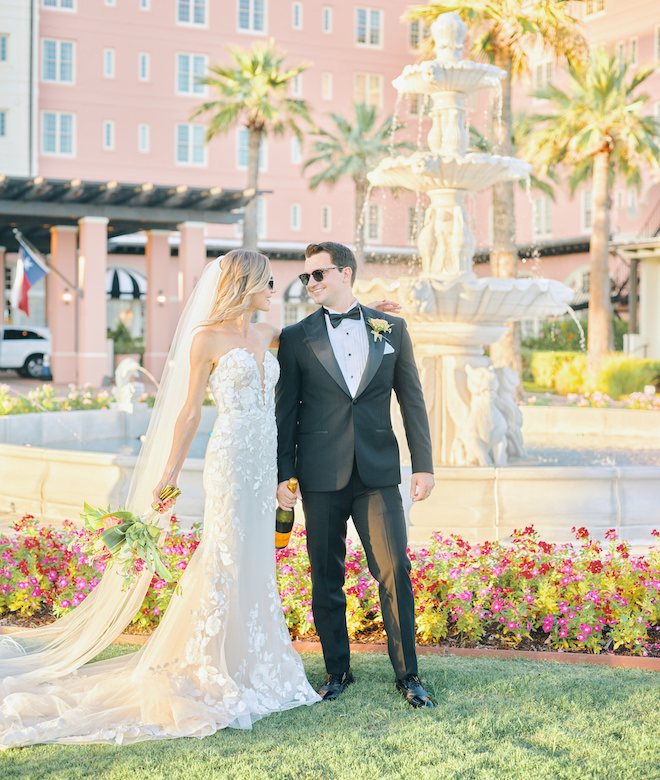A bride and groom smiling at each other while the groom holds a bottle of champagne and the bride holds her bouquet in the center lawn at the Grand Galvez.