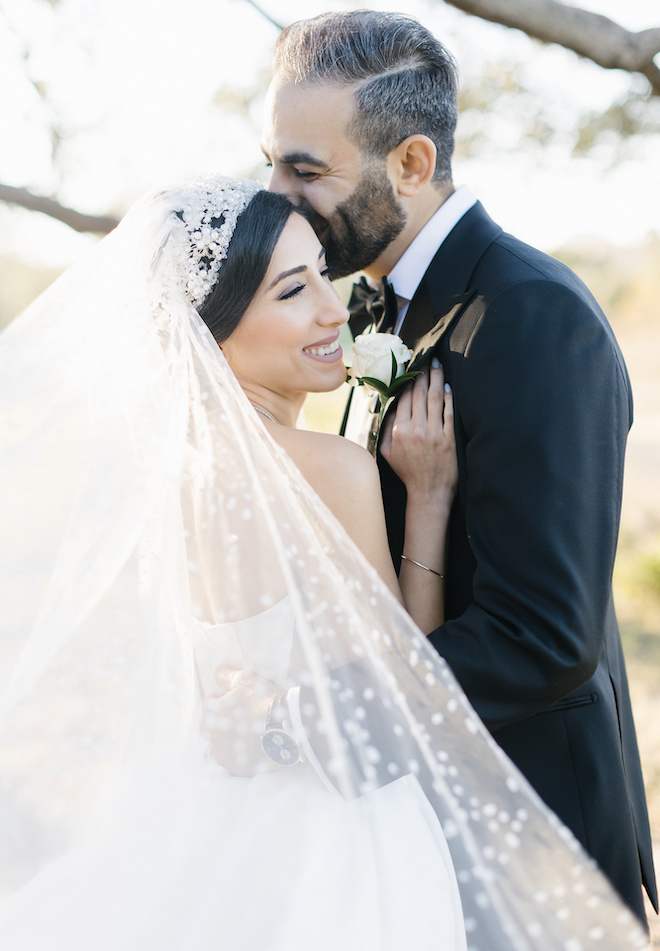 The groom kissing the brides head as she smiles and her veil flows.