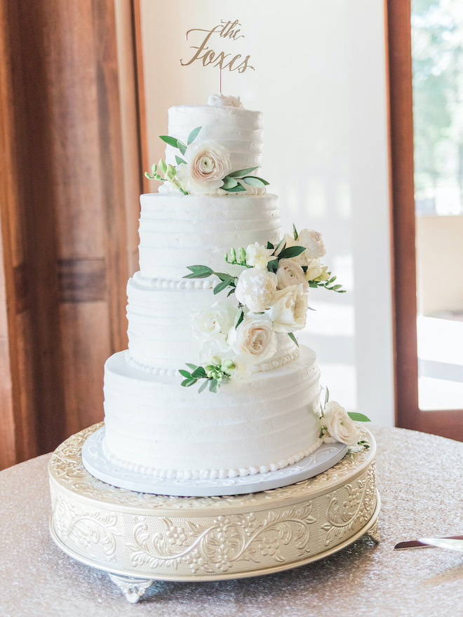 A four tier white wedding cake with a "The Foxes" sign and sage and blush florals. 