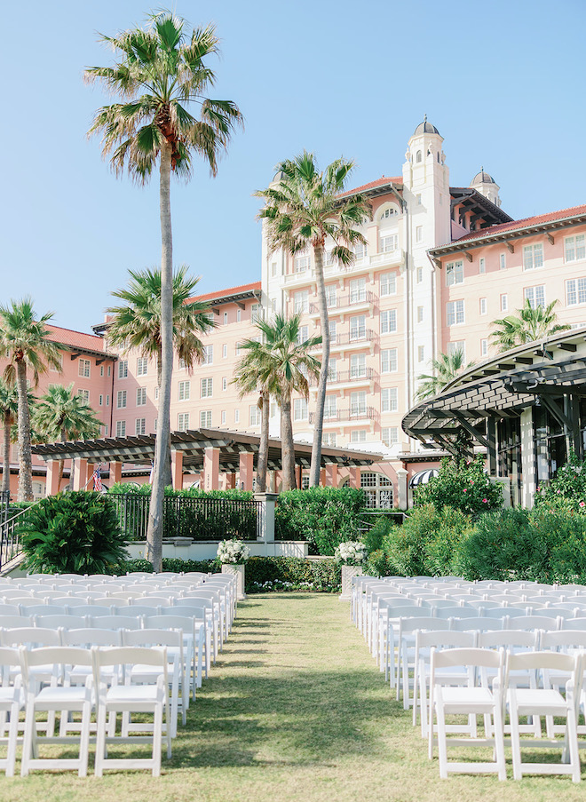 White chairs set up for a ceremony on the lawn in front of the pink hotel with palm trees and green landscaping. 