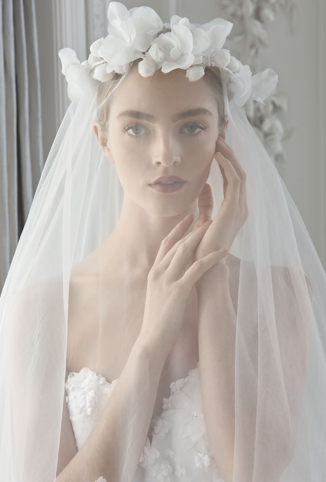 A woman touching her cheek with a white flower crown veil over her face.