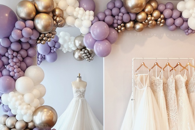 Purple, silver and gold balloons decorating the bridal shop with wedding dresses on a mannequin and a rack.