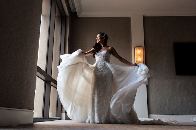 The bride looking out the window while picking up the sides of her dress. 