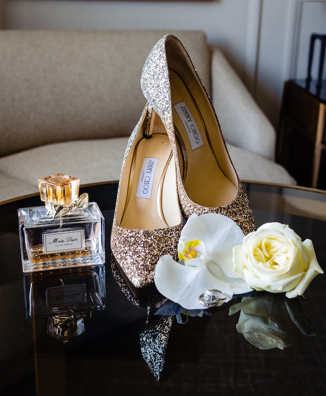 Sparkly Jimmy Choo shoes, Dior perfume and two white flowers on a black table. 