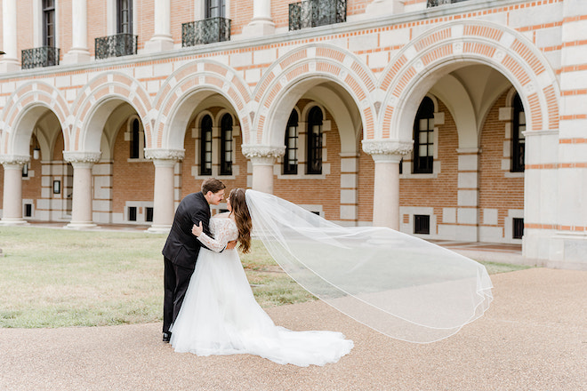 The bride and groom about to kiss with her veil flowing in the wind in front of Rice University. 