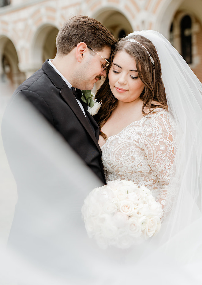 A groom resting his head on the brides cheek as her veil flows in the wind and she is holding a bouquet of white flowers. 