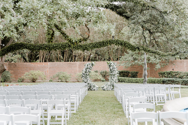 The ceremony setup with a broken arch and white chairs under a large green tree. 