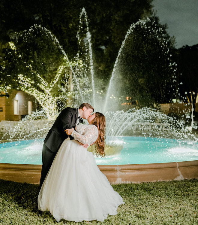 A bride and groom kissing in front of a large fountain.